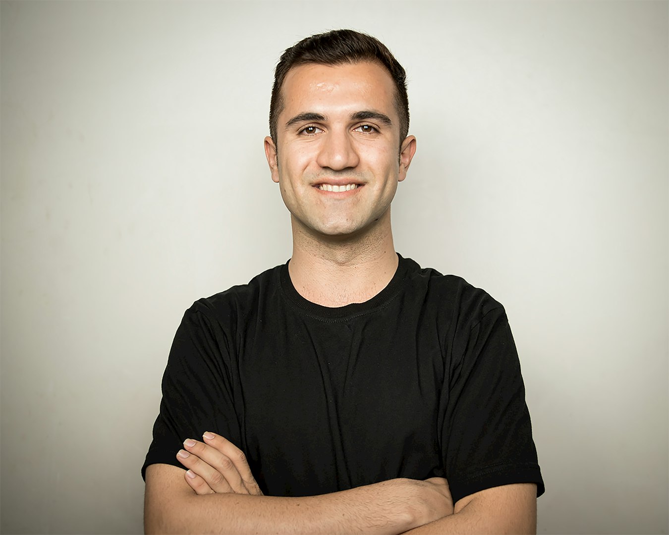 A man with wavy brown hair and a beard is standing in front of a shelf filled with various household items. He is wearing a light blue polo shirt and has a neutral expression.